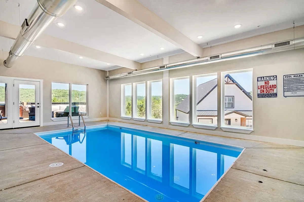 The image showcases a stunning indoor pool framed by expansive windows that reveal tranquil views of the surrounding natural landscape. Soft natural light pours into the space, creating an inviting, relaxing ambiance perfect for swimming, lounging, or socializing. The spacious pool area hints at luxury and exclusivity, complemented by upscale finishes and ample room for group gatherings.