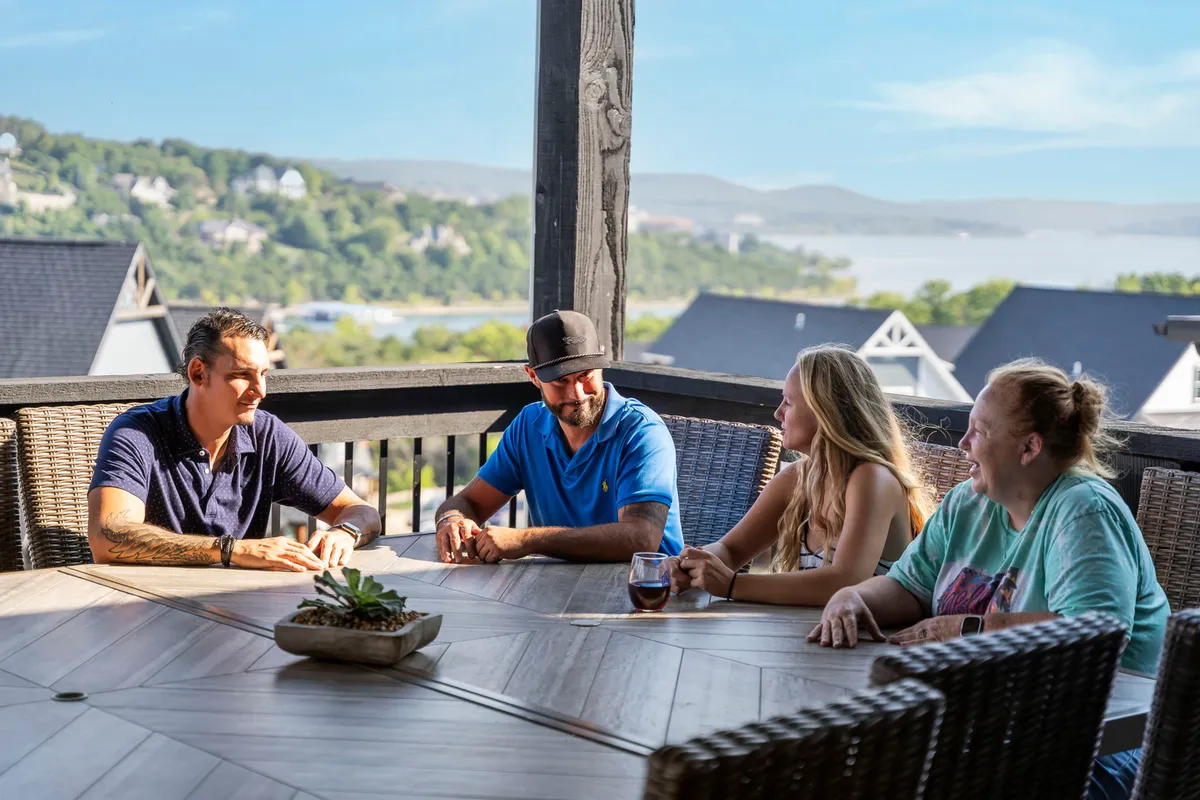 An outdoor dining scene with a group of people seated around a large table, enjoying casual refreshments with panoramic views of hills and water in the background. The atmosphere is relaxed and social, with a decorative succulent adding a touch of style to the setting.