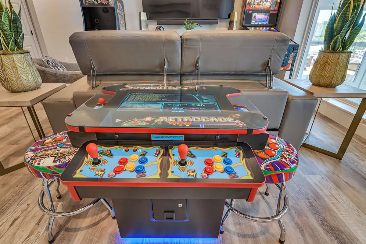 A vibrant interior scene with a central arcade game table, colorful bar stools, lush green plants, and a cozy sofa. The space feels fun and nostalgic, inviting playful competition and group gatherings inside.