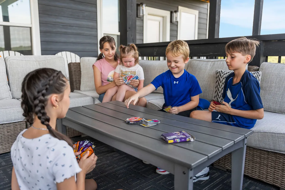 Children play cards at an outdoor table surrounded by comfortable seating, with sunlight and fresh air creating a joyful, relaxed mood. The scene is set in an inviting patio area that encourages family bonding and outdoor enjoyment.