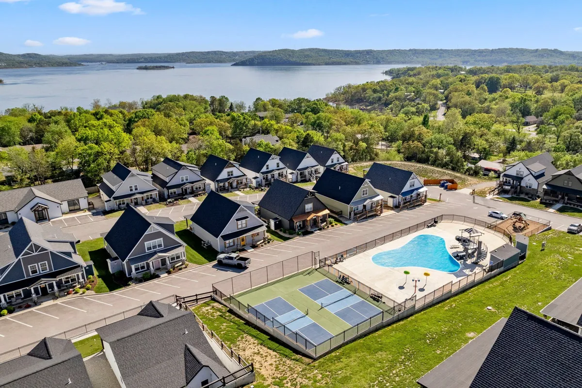 This image features a lively exterior community scene with a swimming pool, tennis courts, residential homes, and a view of the lake in the background. People can be seen engaging in activities such as swimming, playing tennis, socializing, and enjoying the lakeside environment.