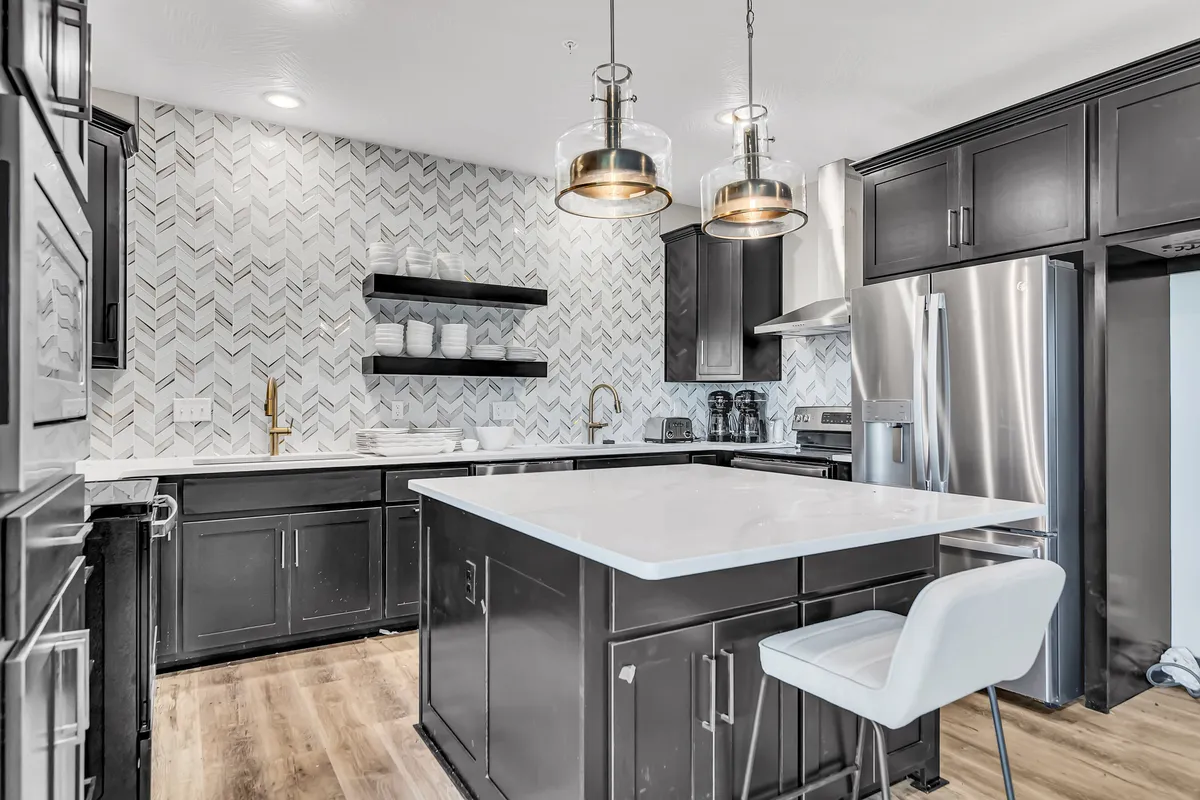 A modern kitchen scene with a central island, premium appliances, shelves with dishware, and contemporary lighting. The open design allows for guests to gather around while meals are being prepared, illustrating a social and functional kitchen environment.